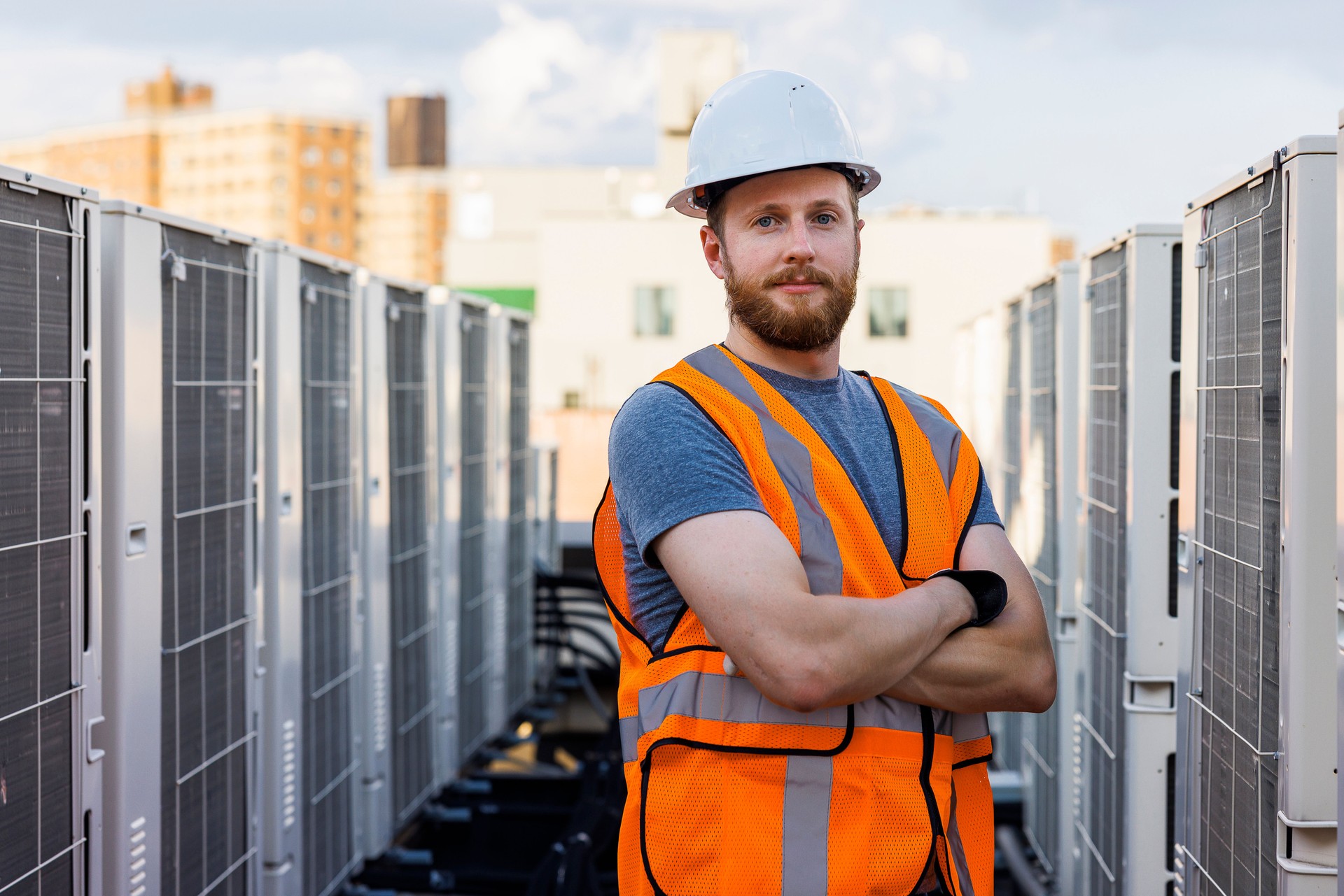 On the roof of the building, a blue collar worker poses near air-conditioning units.
